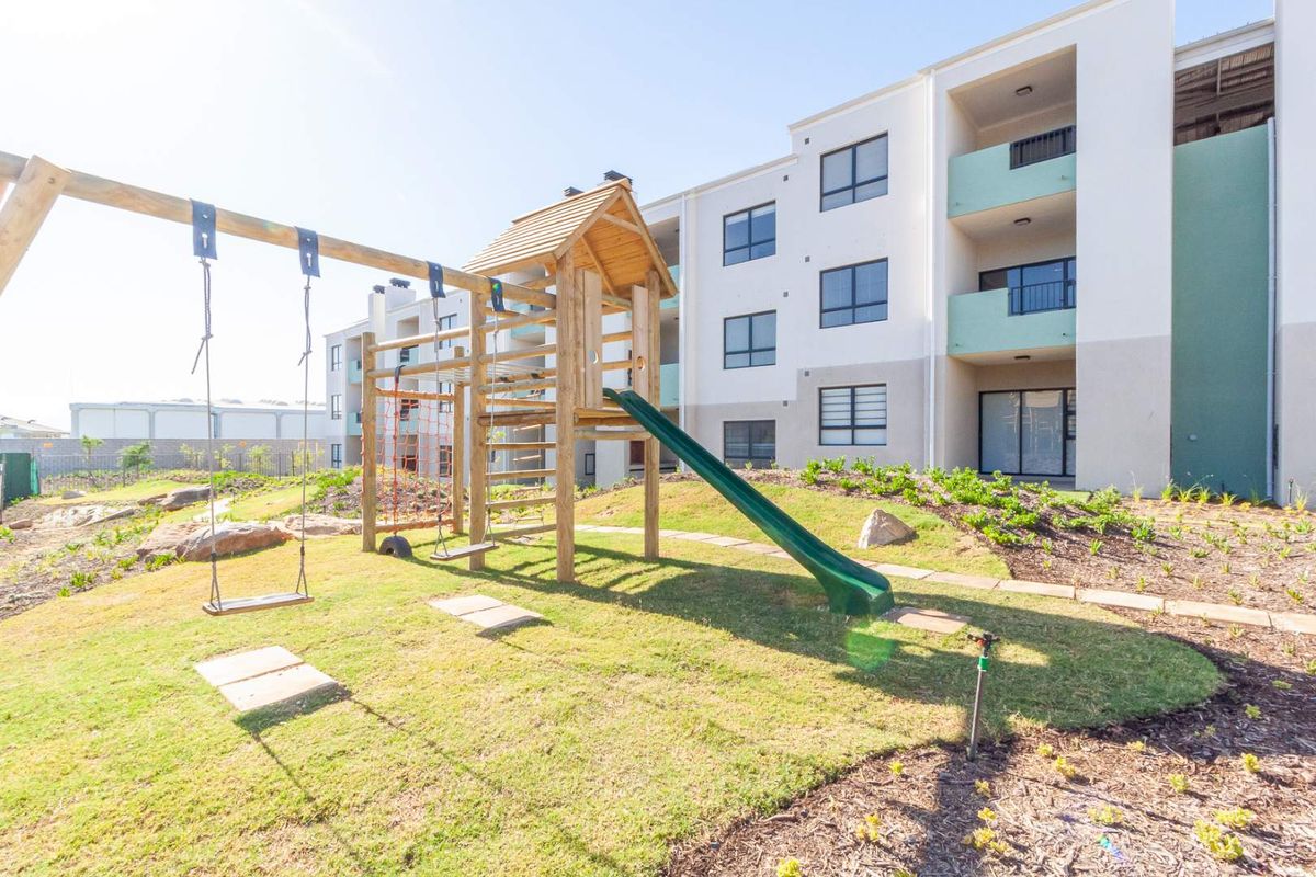 A well-maintained communal garden area featuring a wooden playground set, situated in front of a modern apartment complex with balconies.