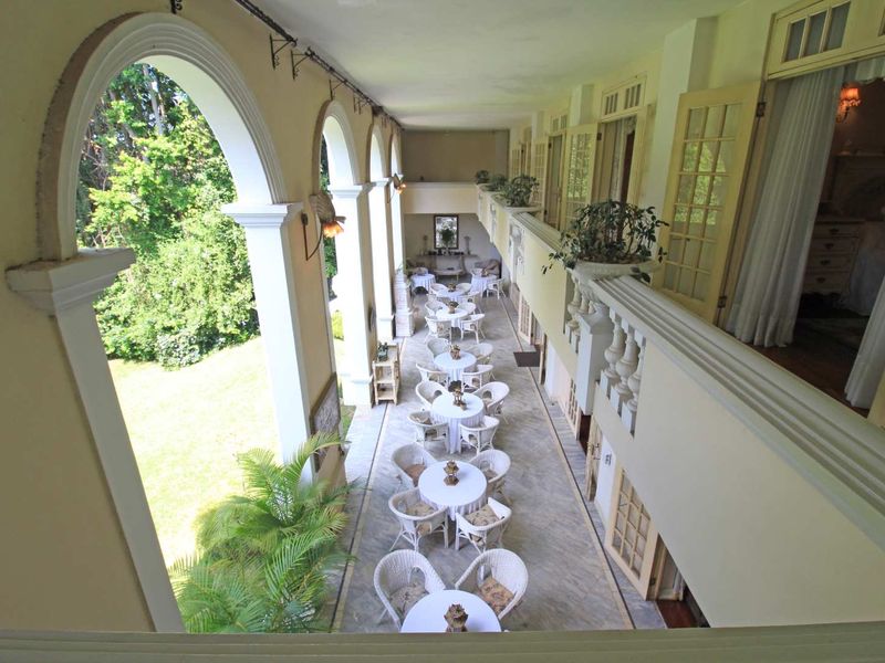 A spacious covered veranda featuring classic arched columns, stone flooring, and a balustrade, providing an elegant outdoor seating area overlooking the garden.
