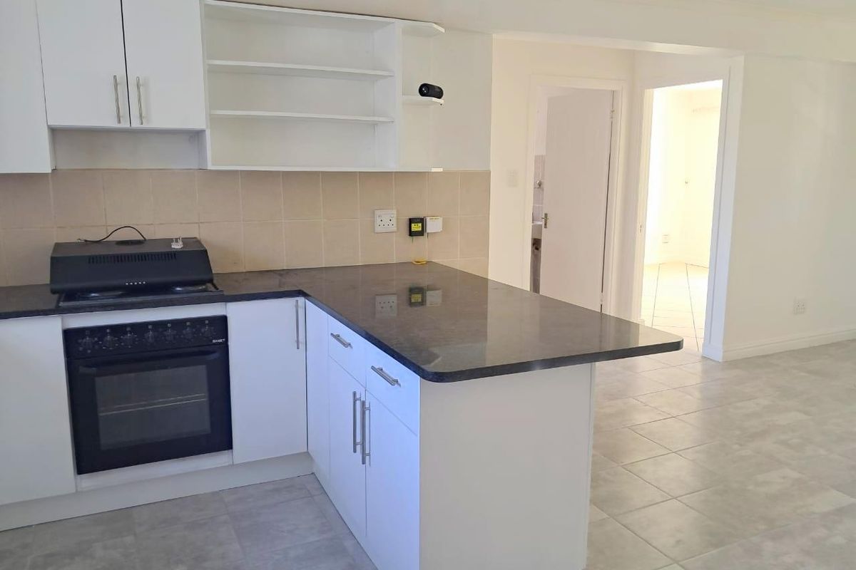 A clean, functional kitchen featuring white cabinetry, dark laminate countertops, a built-in oven, and neutral tiled flooring, leading into an adjacent hallway.