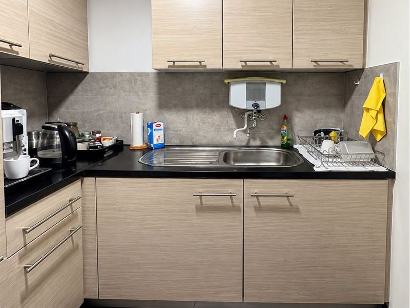 A compact, modern office kitchenette featuring light wood-grain cabinetry, dark stone countertops, a stainless steel sink, and neutral tiled flooring.