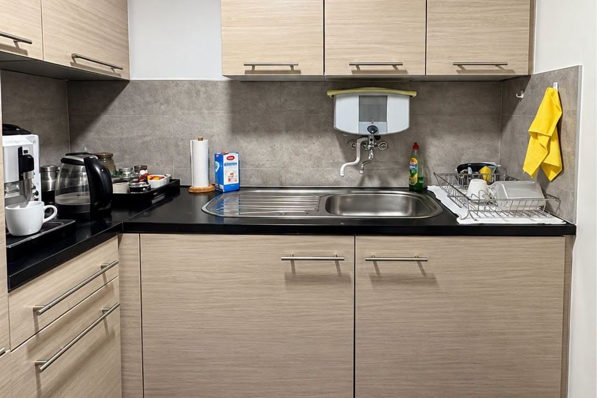 A compact, modern office kitchenette featuring light wood-grain cabinetry, dark stone countertops, a stainless steel sink, and neutral tiled flooring.