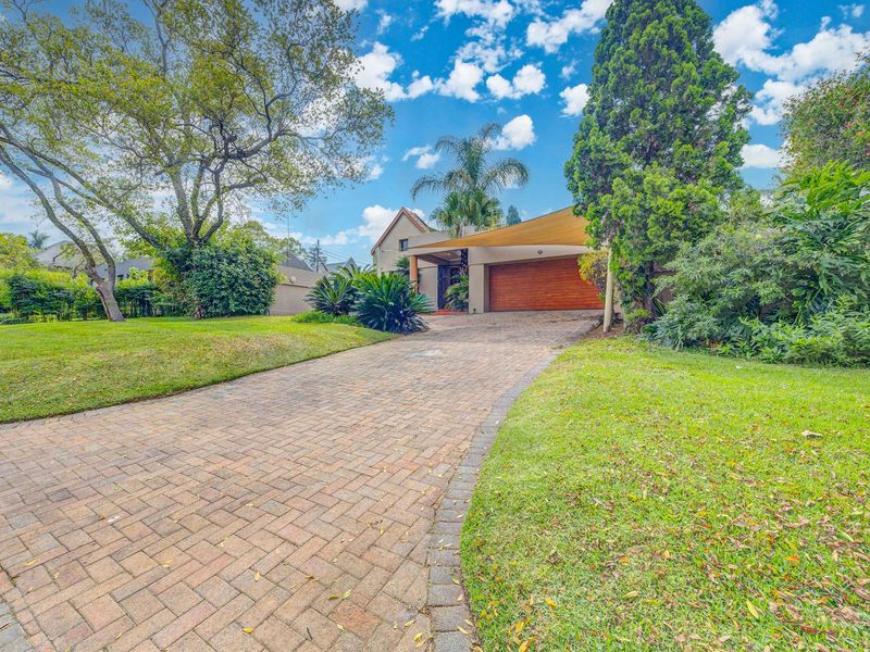 Front view of the property featuring a paved driveway, double garage with wooden door, and a well-maintained landscaped garden with a shade sail.