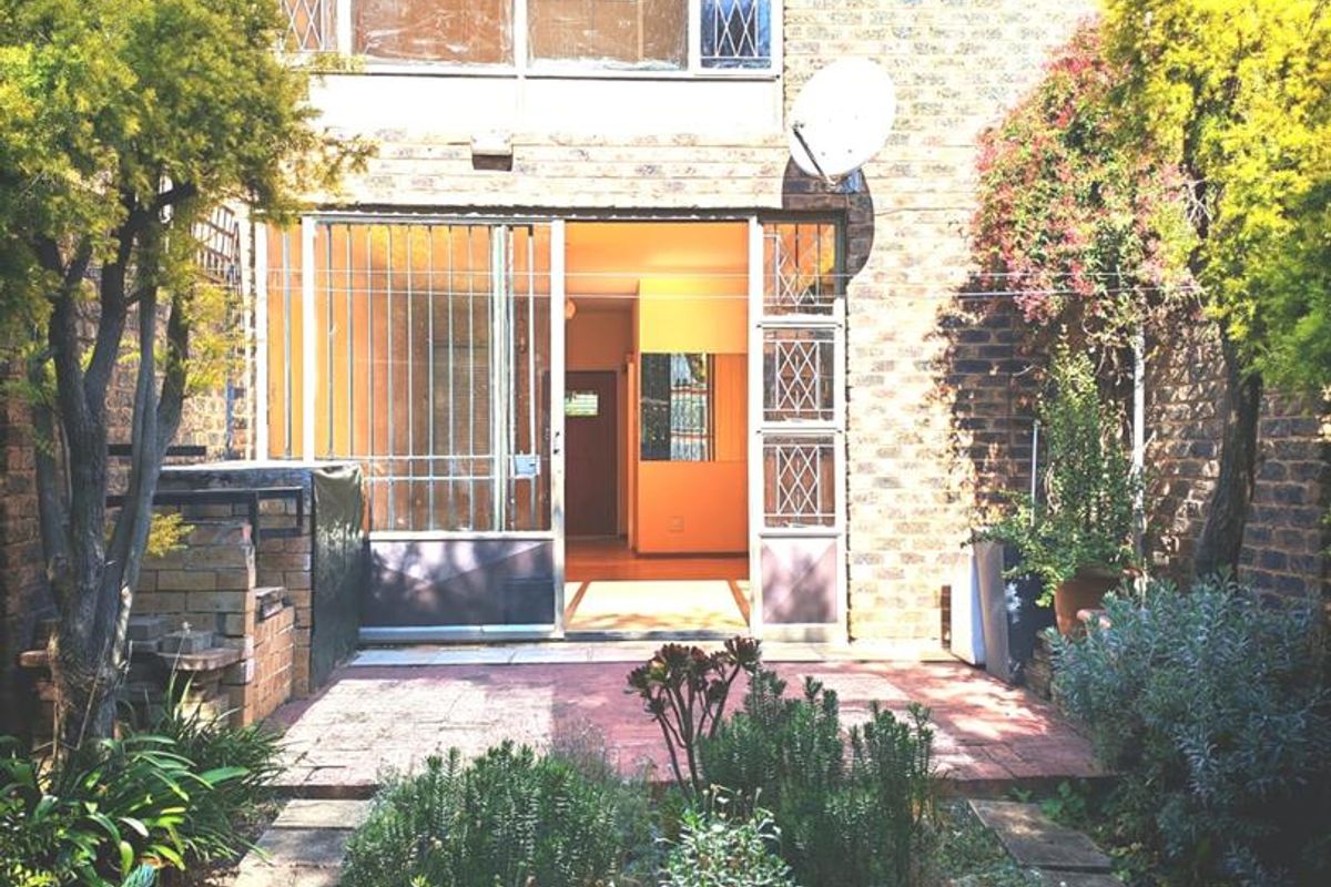 Rear view of the townhouse featuring a paved patio, built-in braai area, and established garden leading to the main entrance.