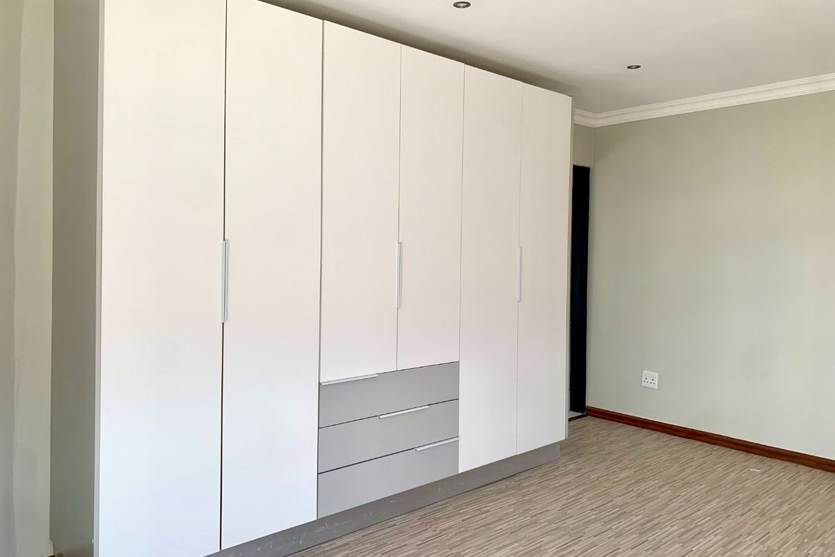 A bright, empty bedroom featuring modern white and grey built-in cupboards, recessed downlights, and durable laminate flooring.