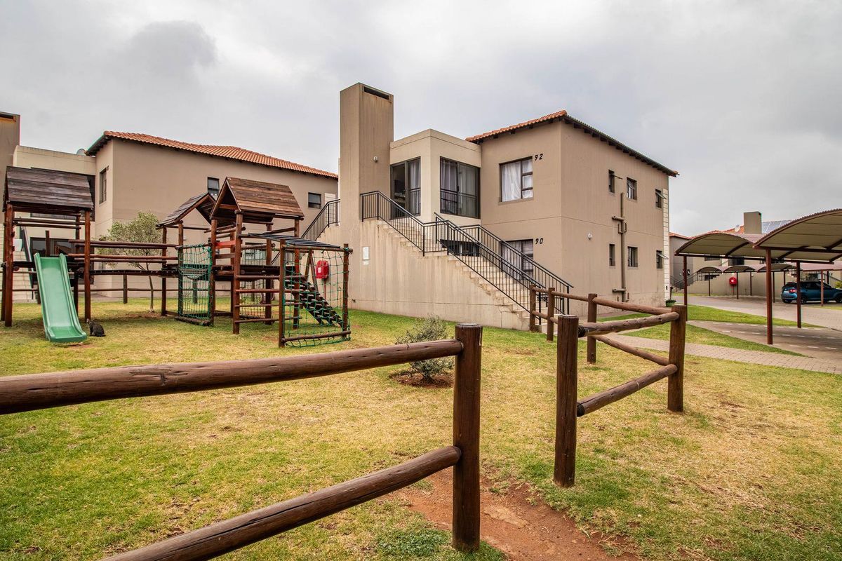 Exterior view of an apartment complex featuring a communal garden area with a wooden playground structure and covered parking bays.