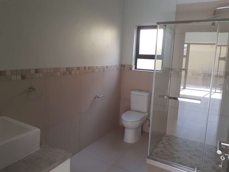 A clean, modern bathroom featuring a glass-enclosed shower, a white toilet, and a vanity basin with neutral wall and floor tiling.