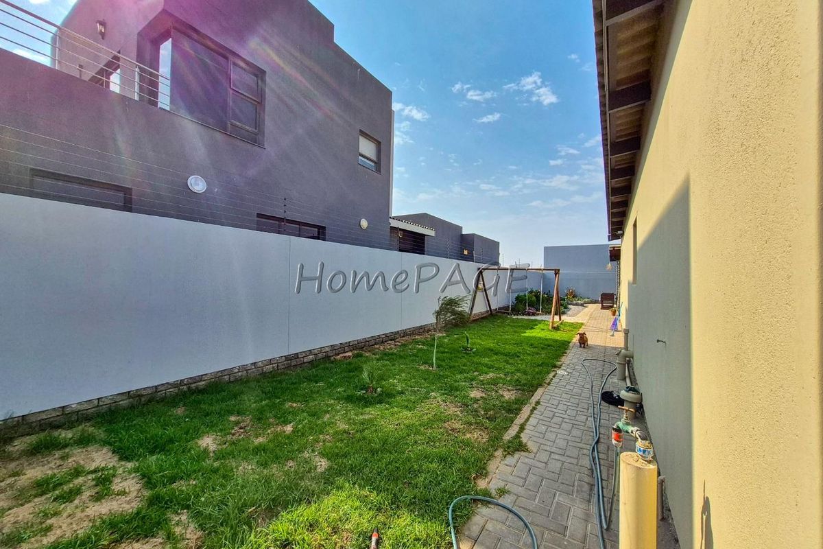 A side garden area featuring a paved walkway, lawn, boundary wall with electric fencing, and modern exterior building architecture.