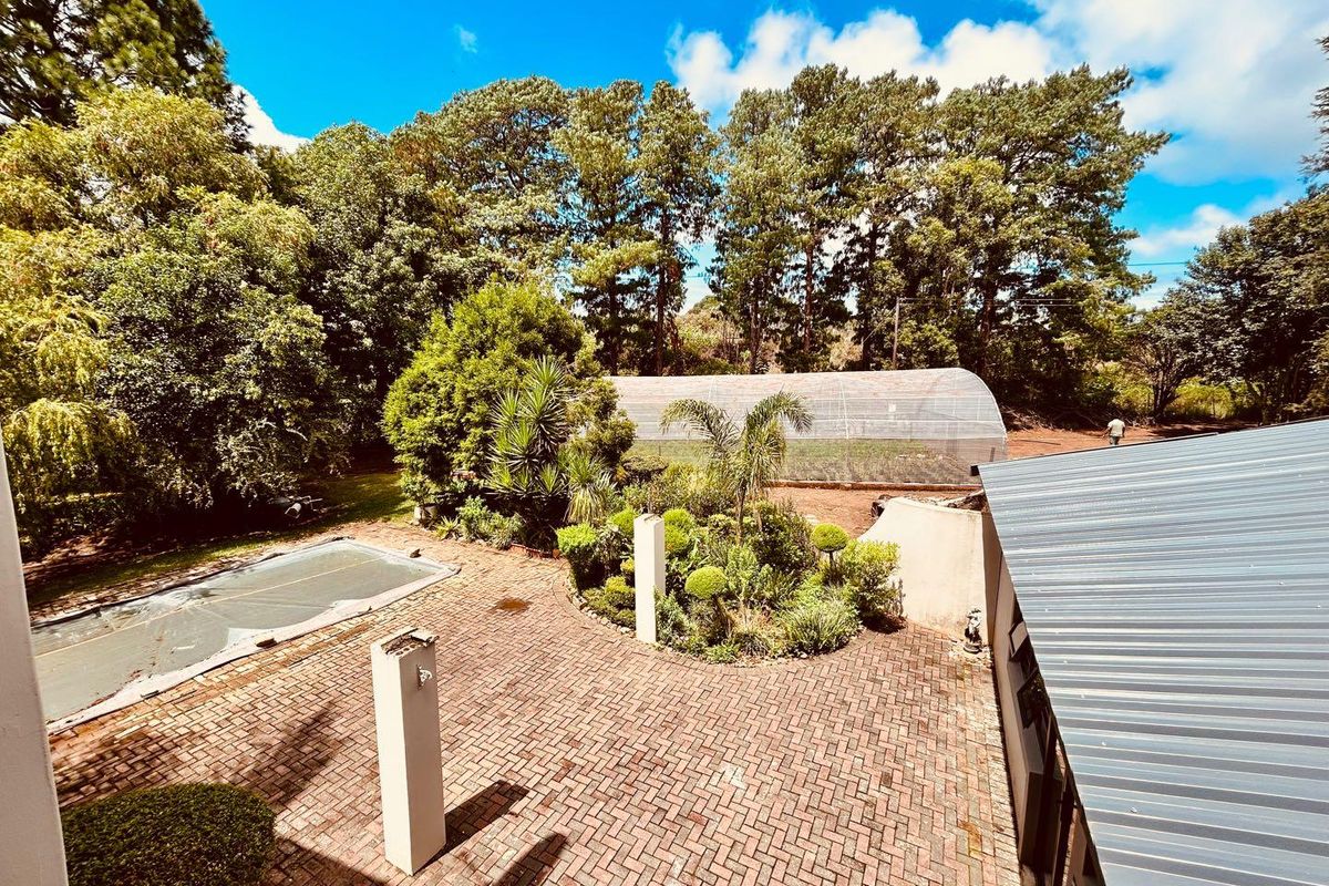 Elevated view of a paved courtyard area featuring a swimming pool with cover, landscaped garden beds, and a large greenhouse structure in the background.