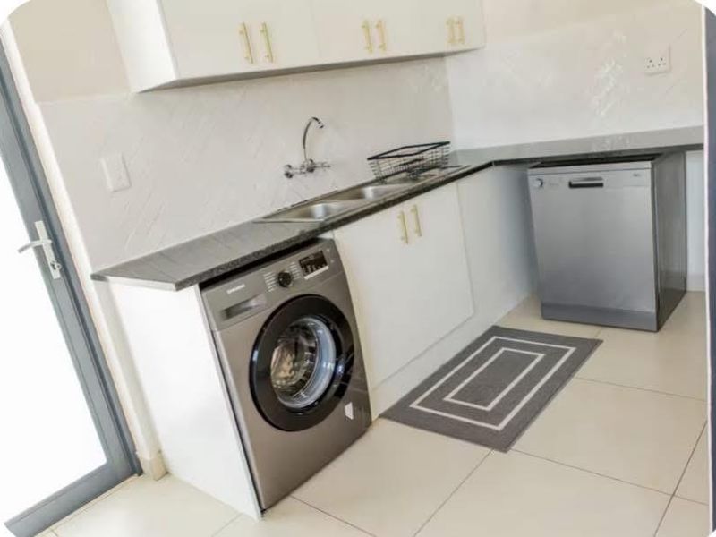 A functional laundry area featuring modern white cabinetry, stone countertops, a double sink, and tiled finishes, providing efficient space for utility tasks.
