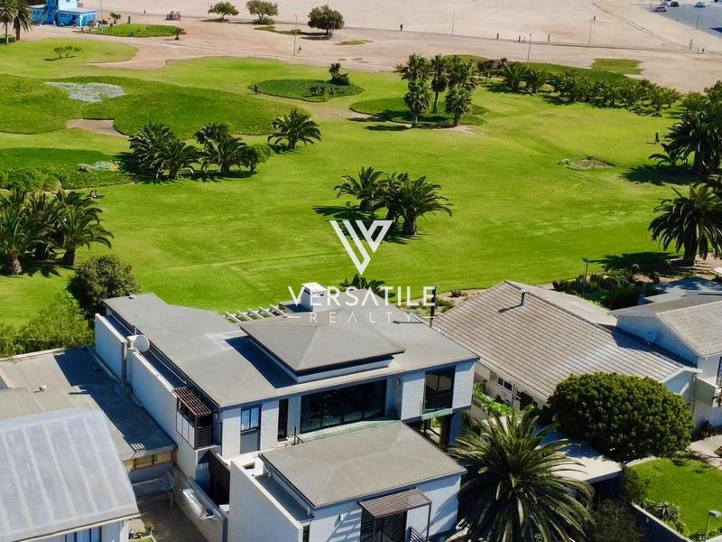 Aerial view of a modern multi-level residence featuring flat roof architecture, balconies, and a prime location adjacent to the beach and coastal parkland.