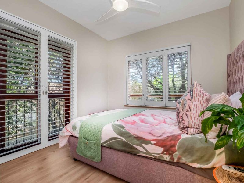 A well-lit bedroom featuring laminate flooring, a ceiling fan, and plantation shutters on both the windows and french doors.