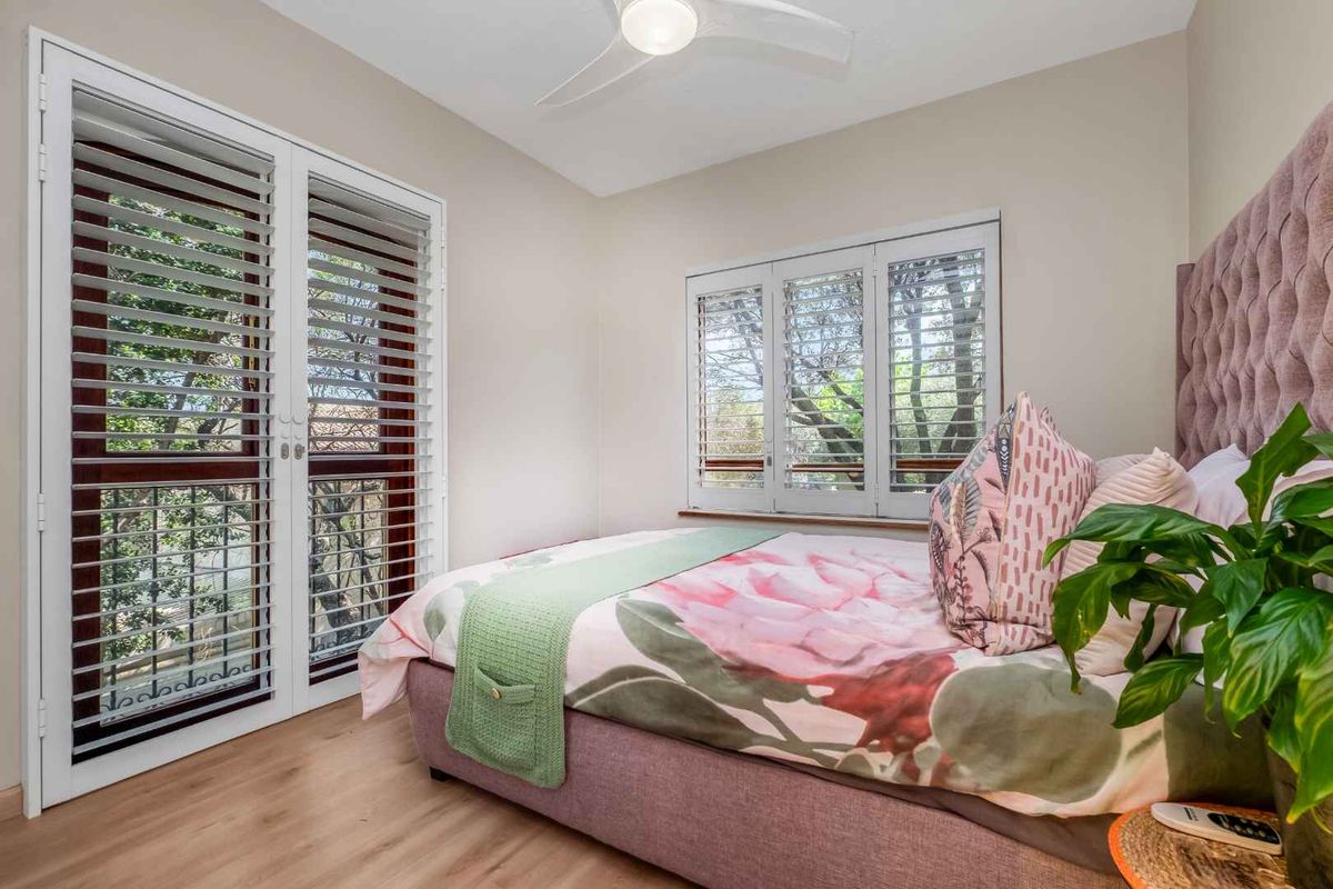 A well-lit bedroom featuring laminate flooring, a ceiling fan, and plantation shutters on both the windows and french doors.