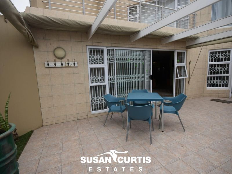 A paved outdoor patio area featuring a pergola, sliding glass doors with security gates, and wall-mounted lighting, suitable for outdoor dining.