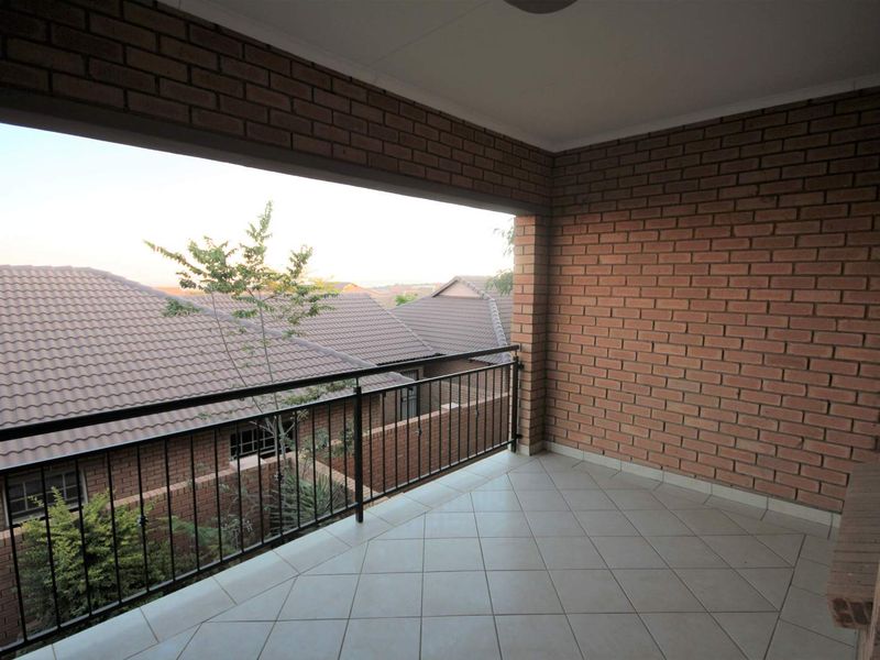 A spacious covered balcony featuring exposed brick walls, tiled flooring, and a metal balustrade, offering an open view of the surrounding residential area.