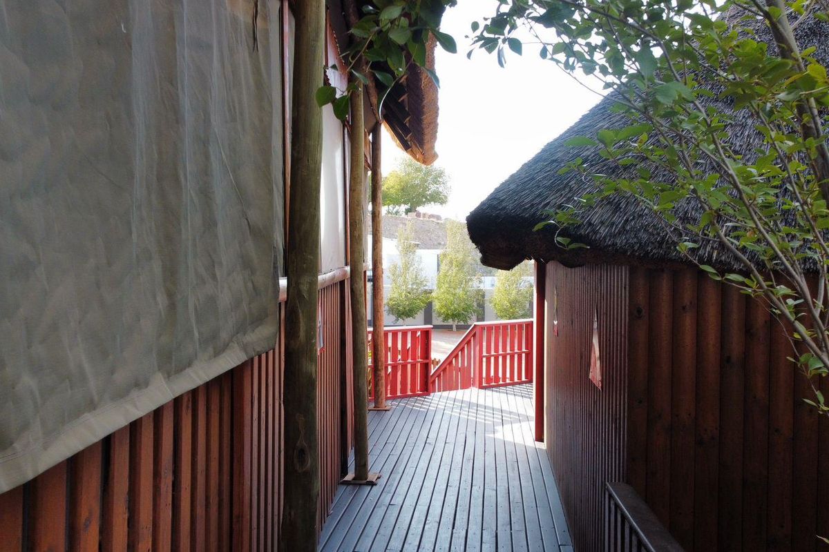 A wooden walkway connecting guest house structures, featuring timber decking, thatched roof overhangs, and red painted railings.