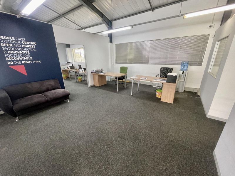Open-plan office space featuring exposed steel roof trusses, carpeted flooring, and large windows with venetian blinds, providing a functional workspace environment.