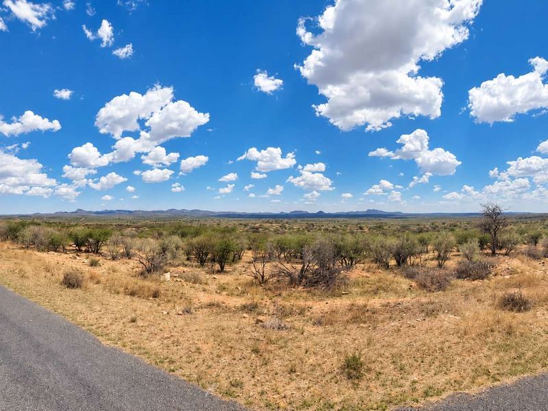 A wide-angle view of a vacant residential plot adjacent to an asphalt road, featuring natural landscape and distant mountain views.
