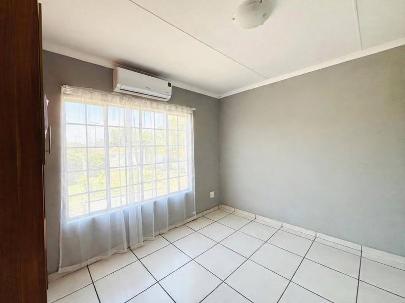 A bright, empty bedroom featuring tiled flooring, a wall-mounted air conditioning unit, and a large window with security bars.