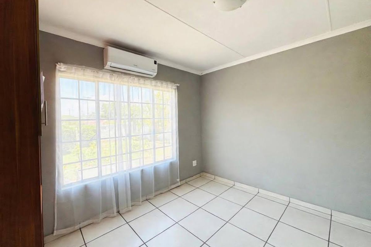 A bright, empty bedroom featuring tiled flooring, a wall-mounted air conditioning unit, and a large window with security bars.