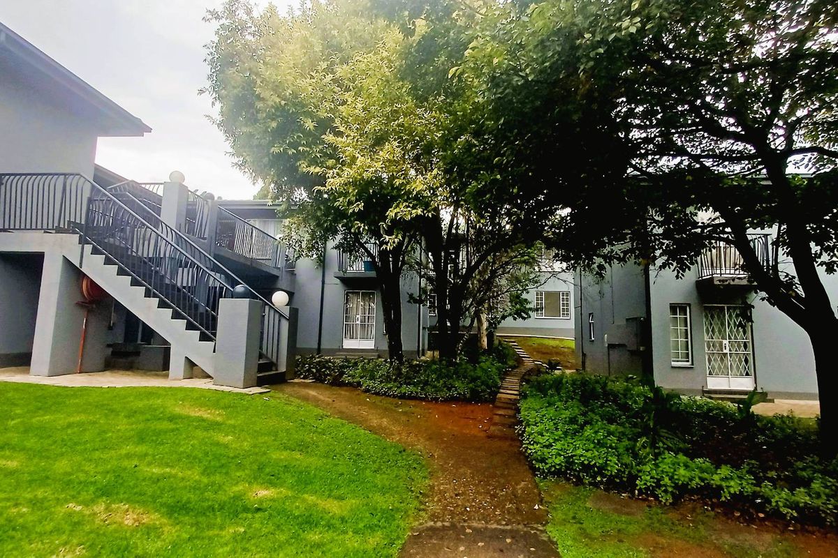 Exterior view of an apartment complex featuring landscaped garden areas, mature trees, and external staircases providing access to upper-level units.