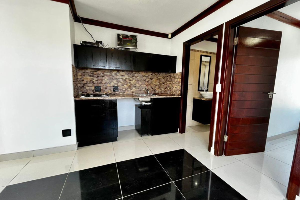 Compact kitchenette featuring dark cabinetry, mosaic tile backsplash, and integrated sink, with an adjacent doorway leading to a bathroom.