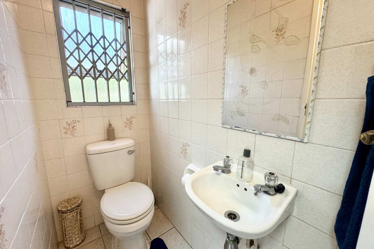 A compact bathroom featuring dated ceramic wall tiles, a pedestal basin, a standard toilet, and a window fitted with a security gate.