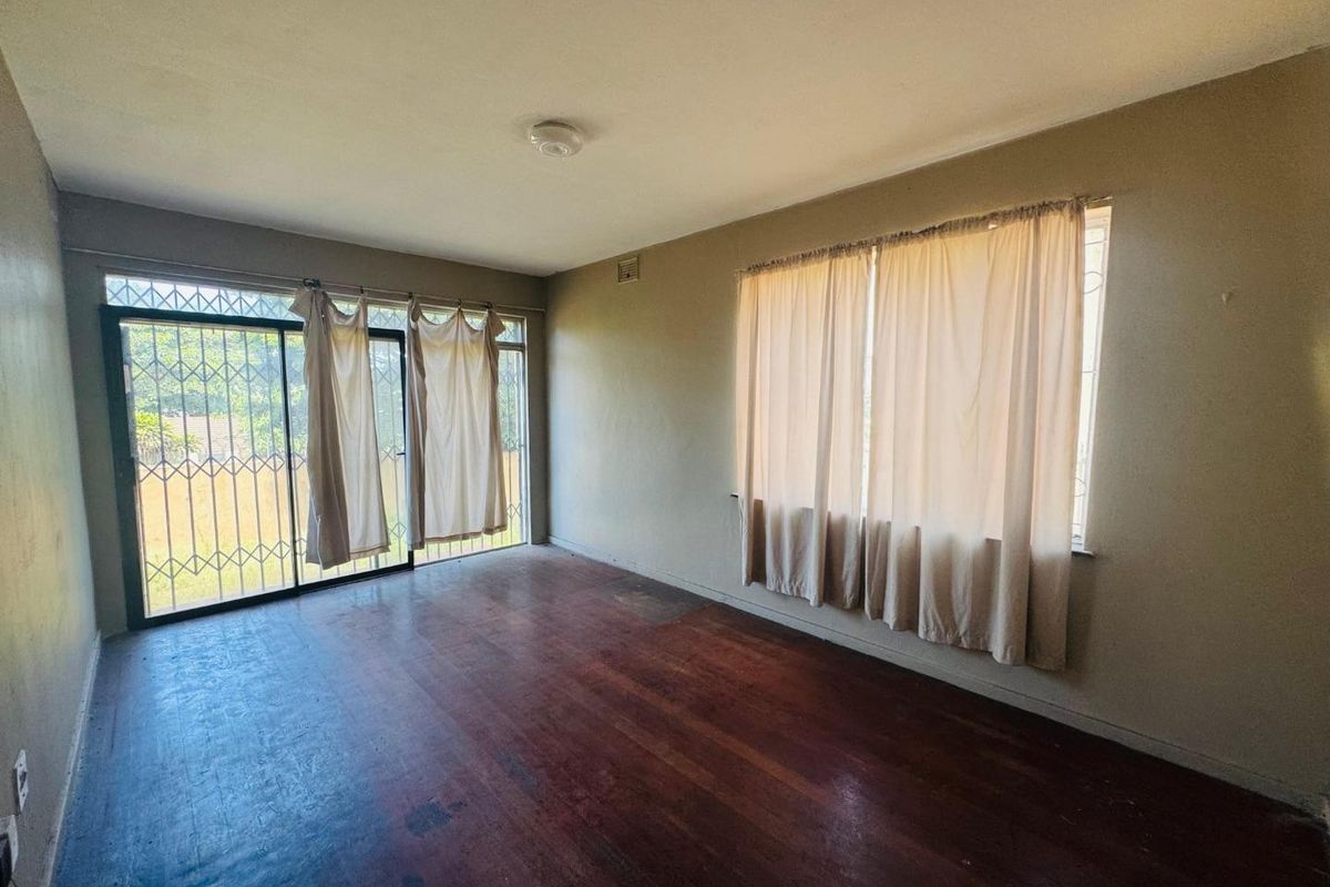 An empty living room featuring original hardwood flooring, large windows, and a sliding glass door with security gate access.