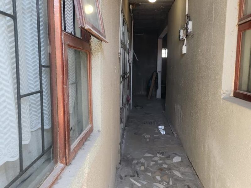 A narrow, unfinished exterior-facing hallway featuring exposed concrete ceilings, timber-framed windows with security bars, and damaged tiled flooring requiring significant repair.