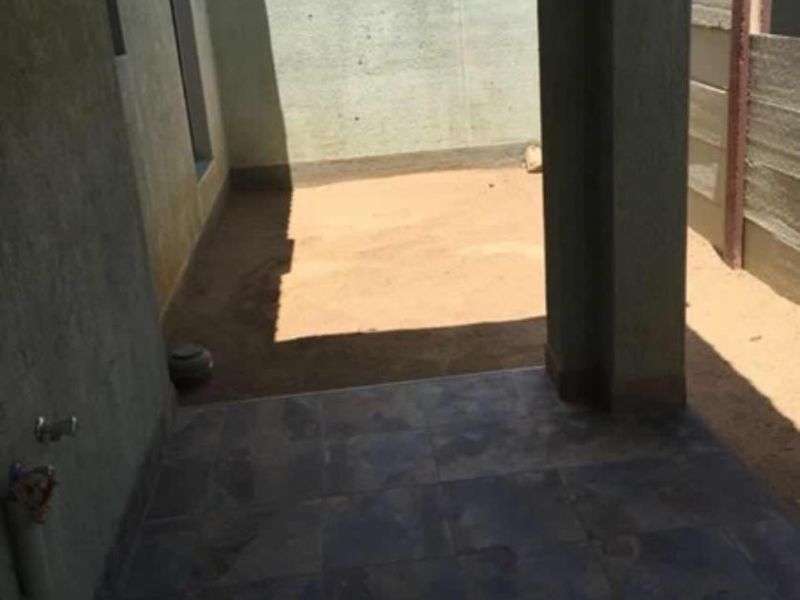 A covered patio area featuring tiled flooring and a concrete support pillar, leading out to an unfinished sandy backyard space.