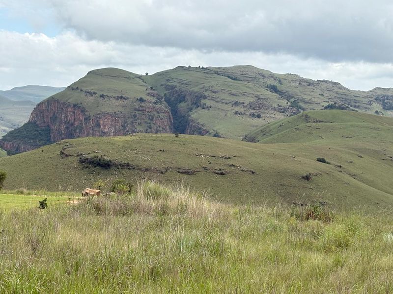 A scenic view of rolling green hills and rocky mountain cliffs under a cloudy sky, showcasing the natural landscape of the vacant land.