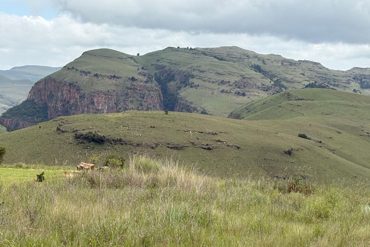 A scenic view of rolling green hills and rocky mountain cliffs under a cloudy sky, showcasing the natural landscape of the vacant land.