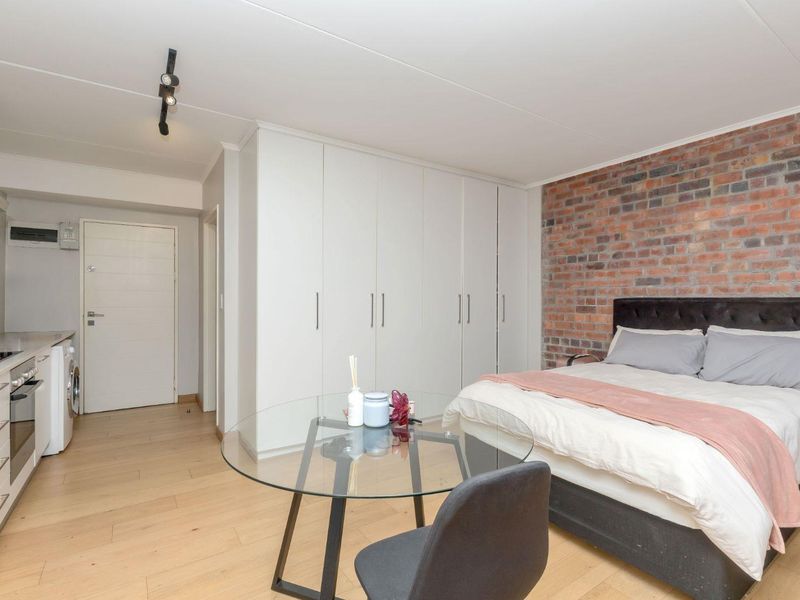 A modern open-plan studio apartment featuring a sleek kitchen, ample built-in cupboard space, and a striking exposed brick feature wall.