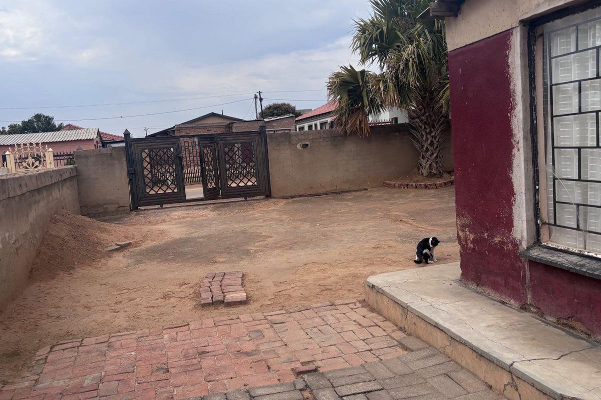 Rear exterior area featuring a paved patio, perimeter wall, and metal security gate, requiring landscaping and exterior maintenance to improve curb appeal.