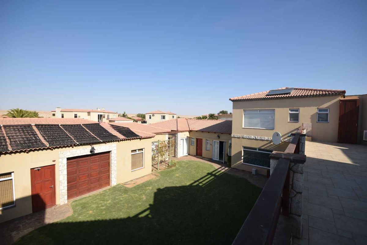 An elevated view of the rear courtyard featuring a central lawn, paved terrace, garage access, and solar water heating panels on the roof.