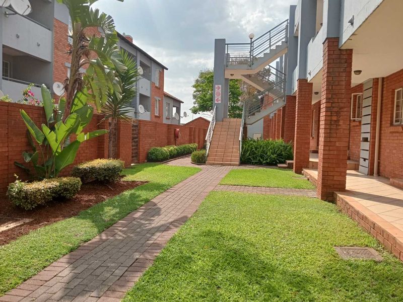 A well-maintained communal garden area featuring a paved walkway, brick building facades, and an external staircase providing access to upper-level apartment units.