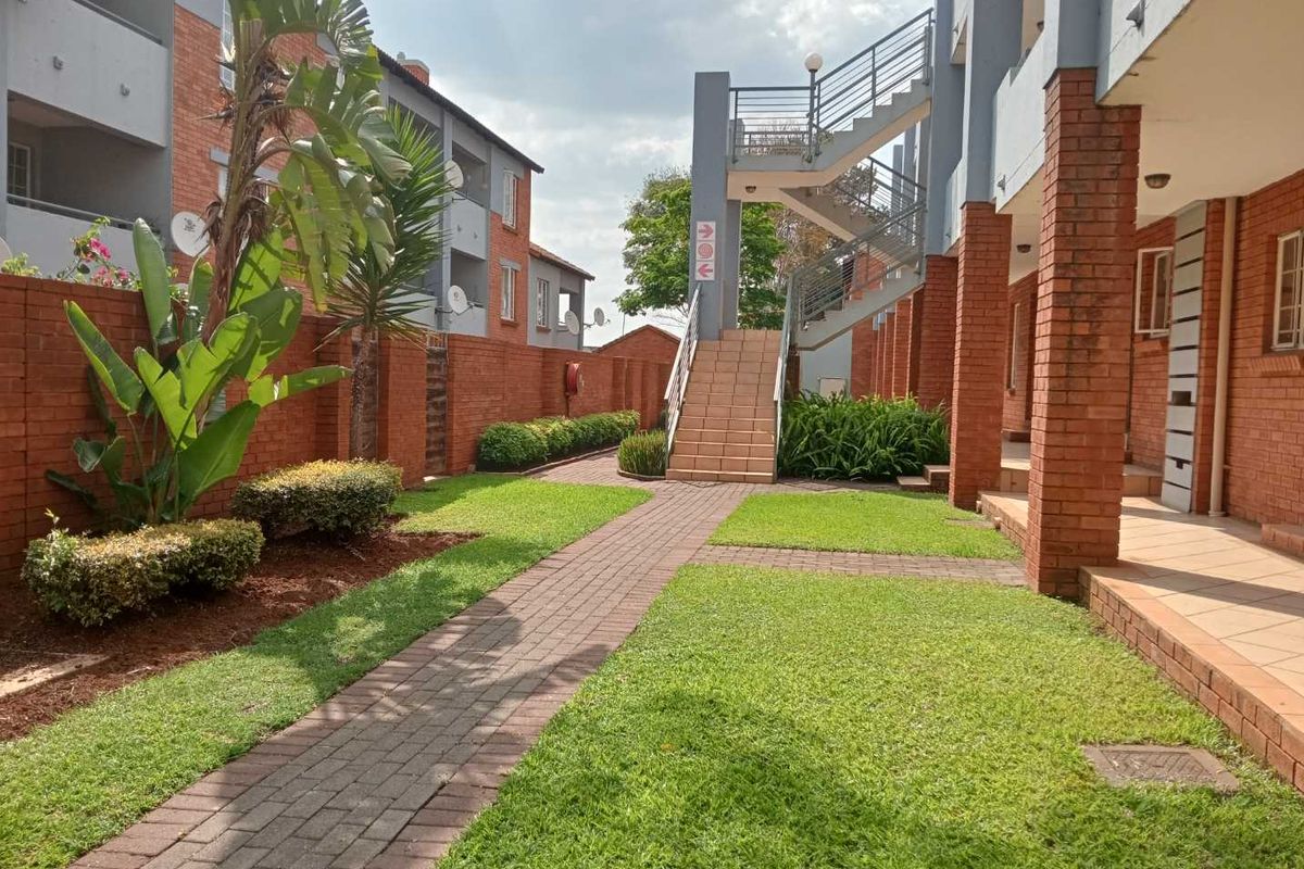 A well-maintained communal garden area featuring a paved walkway, brick building facades, and an external staircase providing access to upper-level apartment units.