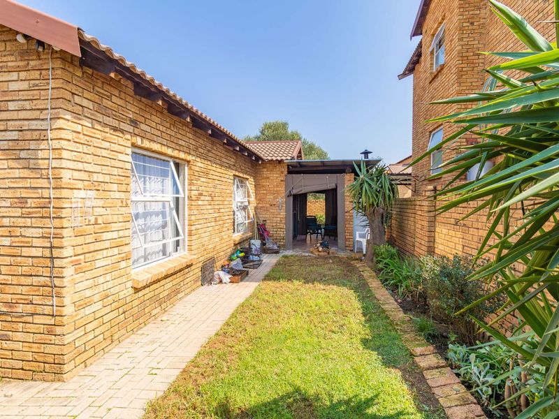 A rear exterior view of a face brick townhouse featuring a paved walkway, a small lawn area, and a covered patio space.