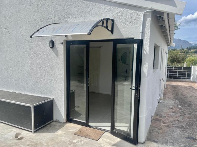 A clean exterior rear view featuring a modern polycarbonate door awning, black aluminium framed glass double doors, and a paved patio area.