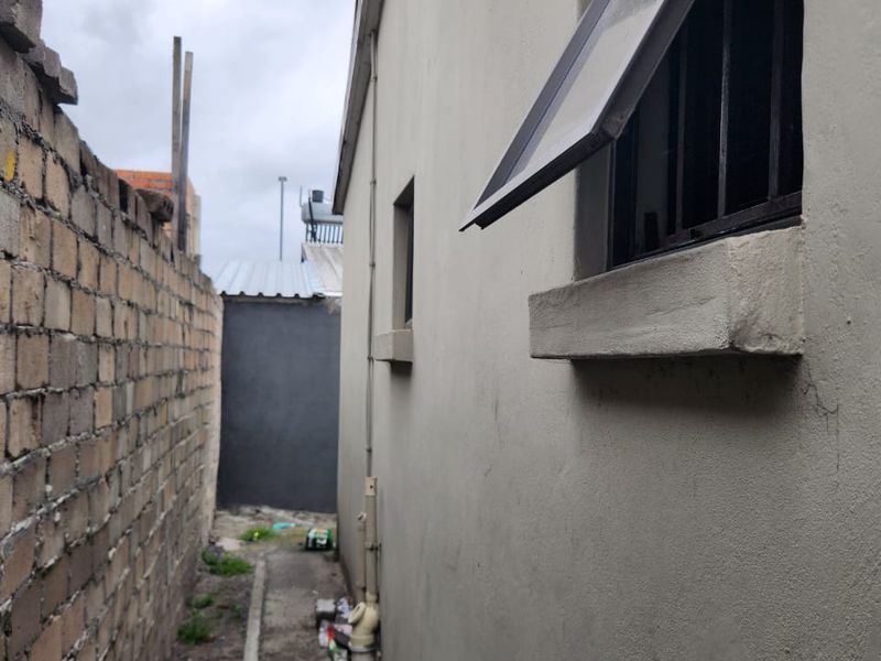 A narrow side passage between the house exterior and a brick boundary wall, featuring security-barred windows and exposed external plumbing.