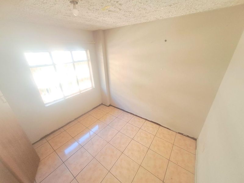 An empty bedroom featuring ceramic floor tiles, a textured ceiling, and an aluminium window frame, requiring minor cosmetic updates to skirting and walls.