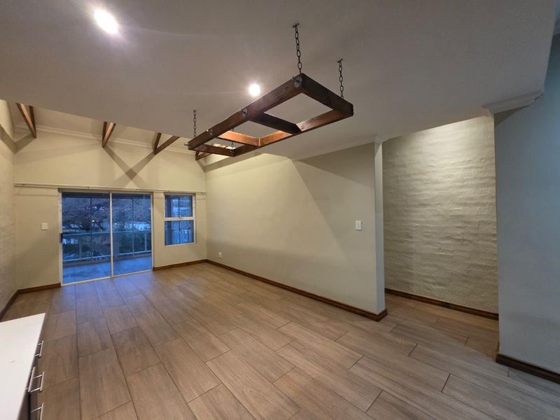 Spacious open-plan living area featuring exposed timber beams, a painted brick feature wall, modern wood-look tile flooring, and access to a private balcony.
