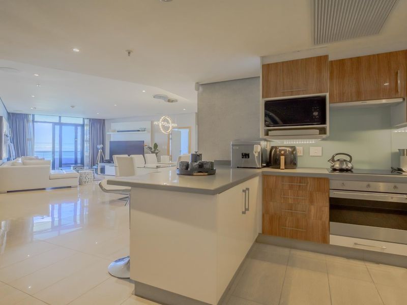 Modern open-plan apartment featuring a sleek kitchen with wood-finish cabinetry, stone countertops, and integrated appliances, flowing into a bright living area with balcony access.