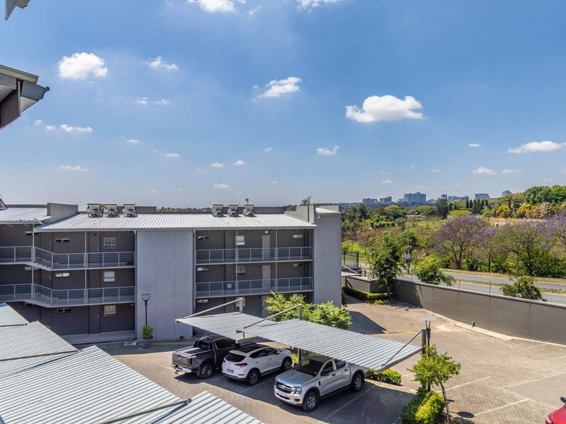 Elevated view of the apartment complex exterior, featuring covered parking bays, paved driveway, and surrounding landscaped grounds under a clear sky.