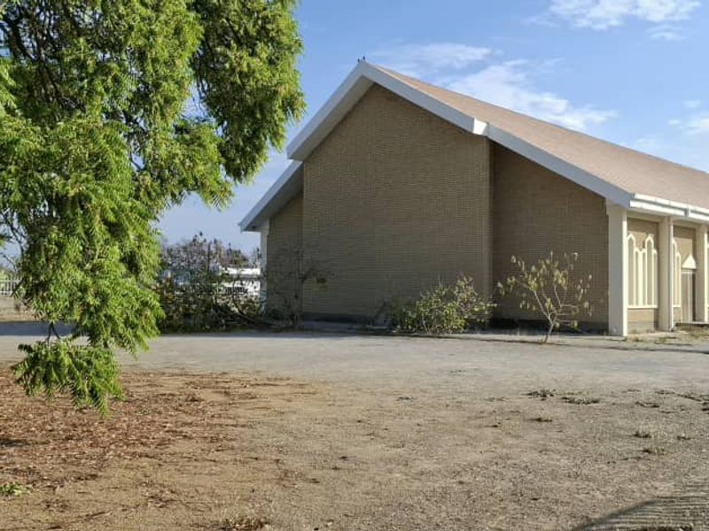 Exterior view of a brick building featuring a pitched roof and arched windows, situated on a large paved lot with mature trees.