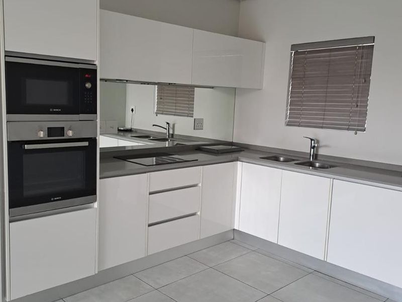 Modern kitchen featuring white cabinetry, built-in oven and microwave, sleek grey countertops, mirrored splashback, and neutral tiled flooring.