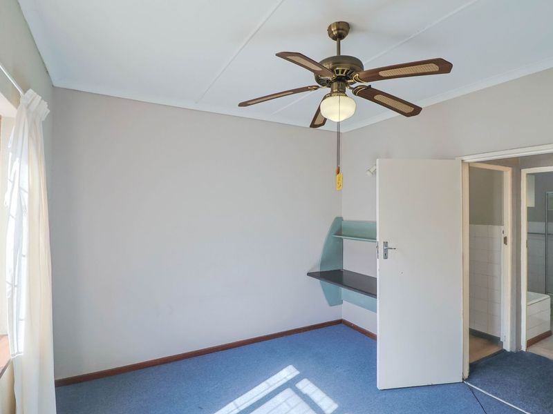 A vacant bedroom featuring a ceiling fan, built-in desk with shelving, and carpet flooring, with an adjacent en-suite bathroom visible.
