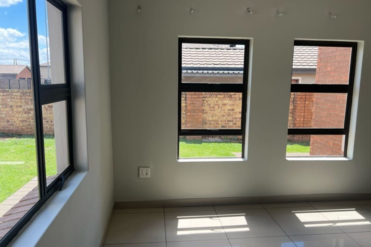 A bright, empty living area featuring modern tiled flooring, neutral walls, and large aluminium-framed windows overlooking the garden.