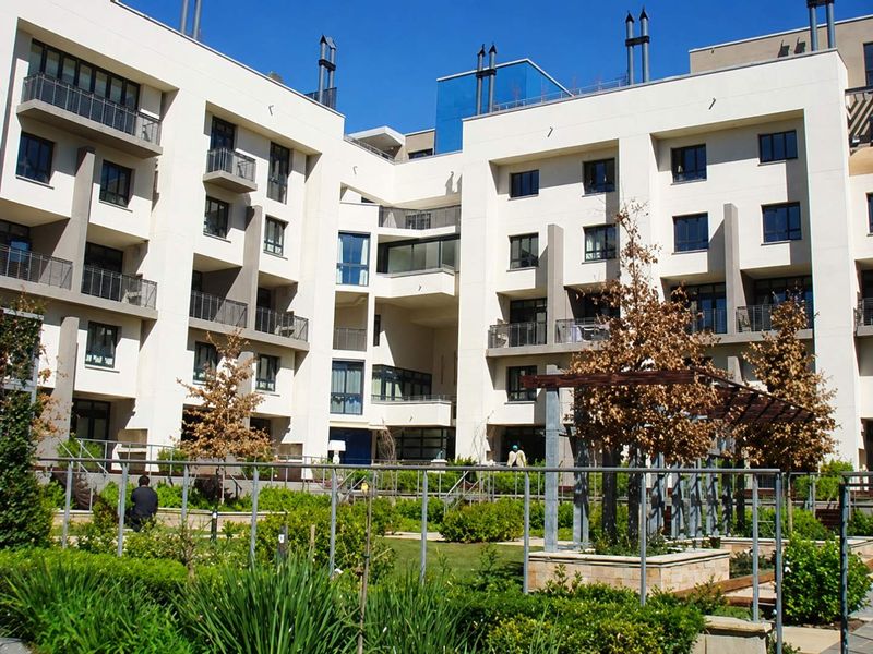 A view of the apartment complex rear facade featuring modern architecture, private balconies, and a well-maintained communal garden area with a pergola.