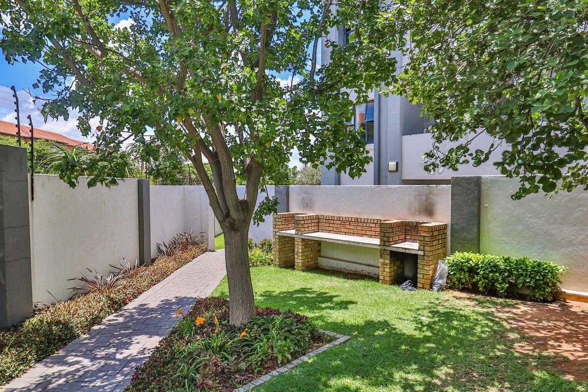 A private rear garden featuring a paved walkway, established tree, and a built-in brick braai area against a perimeter wall.