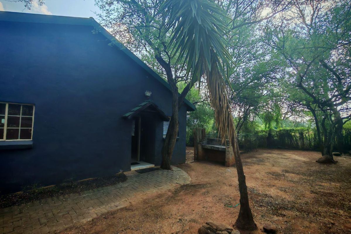 Rear exterior view of the property featuring a paved walkway, built-in braai area, and mature trees in a natural garden setting.